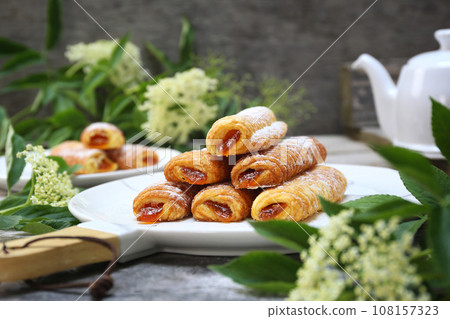 Fripons, French pastries. Apricot jam puff pastry rolls for summer tea and white flowers Fripons, French pastries. Apricot jam puff pastry rolls for summer tea and white flowers 108157323