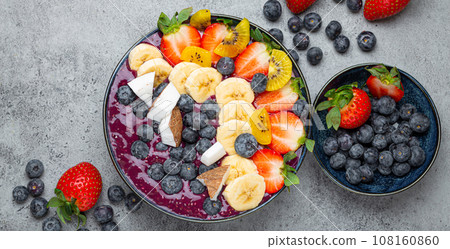 Healthy summer acai smoothie bowl with chia seeds, fresh banana, strawberry, blueberry, cocos, kiwi top view, rustic concrete background with spoon 108160860