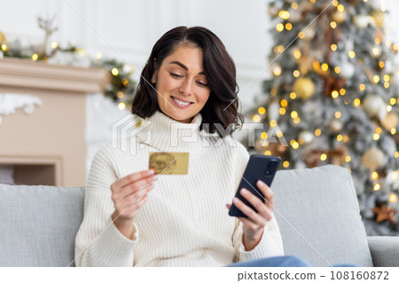 A young beautiful woman is sitting on the sofa at home, holding a credit card and phone and doing online shopping on the eve of the New Year and Christmas holidays. A young beautiful woman is sitting on the sofa at home, holding a credit card and phone and doing online shopping on the eve of the New Year and Christmas holidays. 108160872