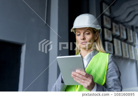 Serious confident thinking female engineer with tablet computer inspecting factory wearing hard hat and reflective vest. 108160895