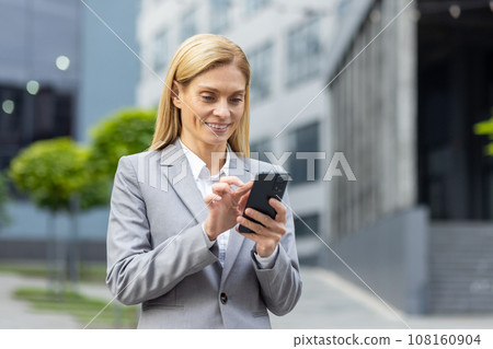 Successful woman boss walks outside office building outdoors, in business suit, businesswoman uses app on phone, smiles contentedly reads internet pages, browses news, writes text message, call. Successful woman boss walks outside office building outdoors, in business suit, businesswoman uses app on phone, smiles contentedly reads internet pages, browses news, writes text message, call. 108160904