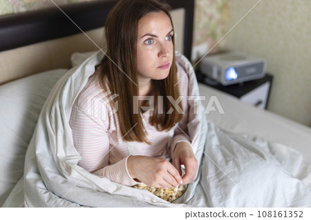 A woman wrapped in blanket watching a movie on her bed using a projector A woman wrapped in blanket watching a movie on her bed using a projector 108161352