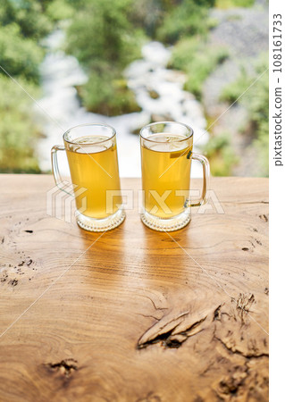 Two cups of ginger root tea with lemon, honey and mint on a wooden table and High-mountain river in the background. Walk in the mountains and relax in a cozy cafe. Hiking on a day off 108161733