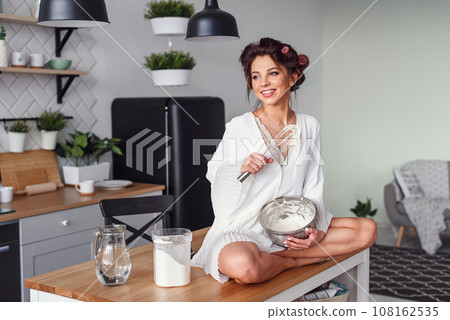 Attractive girl in white bathrobe kneads a dough in a metal plate at stylish modern kitchen. Young pretty woman baking on home kitchen. 108162535
