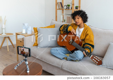 Blogger guitarist. African american girl blogger playing guitar talking to webcam recording vlog. Social media influencer woman streaming at home indoors. Music content creator broadcast tutorial 108163057