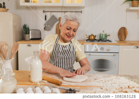 Happy senior woman cooking in kitchen. Stylish older mature gray haired lady grandmother knead dough bake cookies. Old grandma cook homemade food. Household housewife housework concept 108163128