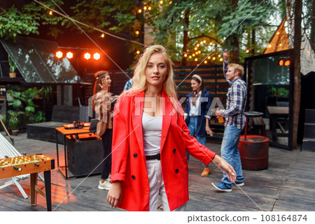 Close up of attractive blond girl with blue eyes posing to the camera with her friends dancing in disco light on the background. Outdoors evening party concept. 108164874