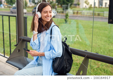 Teen girl listens to the music by white headphones in a public transport station while she waiting for tram. Teen girl listens to the music by white headphones in a public transport station while she waiting for tram. 108164917