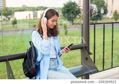 Teen girl listens to the music by white headphones in a public transport station while she waiting for tram. Teen girl listens to the music by white headphones in a public transport station while she waiting for tram. 108164923