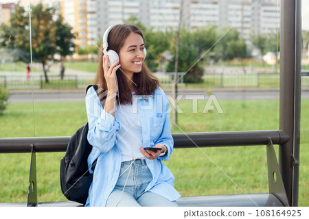 Teen girl listens to the music by white headphones in a public transport station while she waiting for tram. Teen girl listens to the music by white headphones in a public transport station while she waiting for tram. 108164925