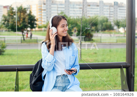 Teen girl listens to the music by white headphones in a public transport station while she waiting for tram. Teen girl listens to the music by white headphones in a public transport station while she waiting for tram. 108164926