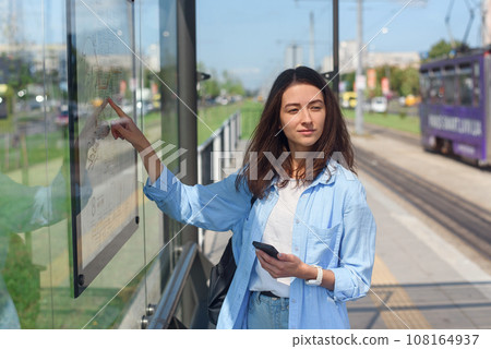 Stylish girl looks to map of public transport to find the right station while standing at the modern tram station outdoors Stylish girl looks to map of public transport to find the right station while standing at the modern tram station outdoors 108164937