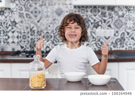 Happy boy feels happiness while eating healthy breakfast of cornflakes and milk at the kitchen. 108164946