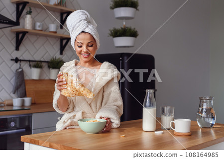 Charming woman in a bathrobe and towel on her head prepares a healthy breakfast with flakes and milk. The girl has a breakfast on stylish cozy kitchen at the morning. 108164985
