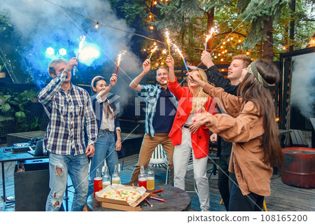 Caucasian friends having garden party outside, they dancing to the Dj's music with bengal lights in house backyard at summer night. 108165200