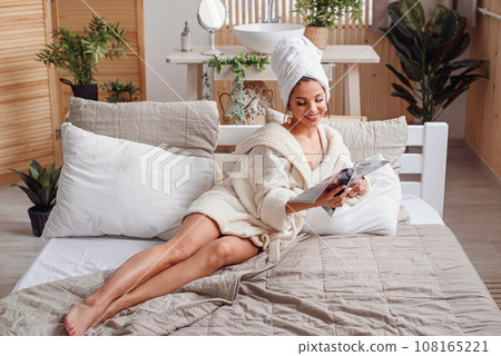 Attractive smiling young woman dressed in white bathrobe lies on the bed at home and reads a new magazine. Beautiful girl enjoying on the bed in morning. Attractive smiling young woman dressed in white bathrobe lies on the bed at home and reads a new magazine. Beautiful girl enjoying on the bed in morning. 108165221