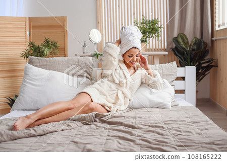 Attractive smiling young woman lies on the bed at the cozy bedroom after morning hygiene procedures dressed in white bathrobe. Beautiful girl having relax on the bed in morning. Attractive smiling young woman lies on the bed at the cozy bedroom after morning hygiene procedures dressed in white bathrobe. Beautiful girl having relax on the bed in morning. 108165222