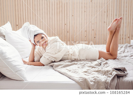 Pretty happy young woman on white bathrobe and with towel on her head lies on the bed at cozy home. Beautiful girl having relax on the bed in morning. 108165284