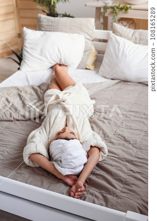 An attractive smiling young woman lying on the bed at home after bath dressed in white bathrobe. Beautiful girl having relax on the bed in morning. 108165289