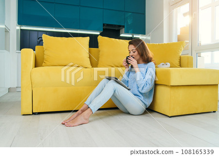 Charming girl holds her tablet computer in hands, sitting on the floor near modern yellow couch and drinks coffee in the modern stylish kitchen. Young cheerful woman having rest at cozy home. Charming girl holds her tablet computer in hands, sitting on the floor near modern yellow couch and drinks coffee in the modern stylish kitchen. Young cheerful woman having rest at cozy home. 108165339