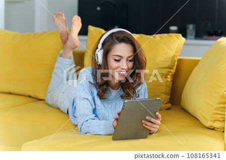 Exuberant girl uses tablet pc while laying on the comfortable yellow sofa and enjoying nice melodies with earphones. 108165341