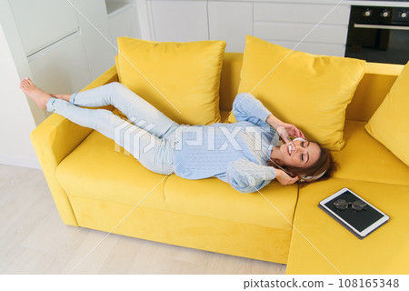 Pretty blonde teenage girl in headphones is listening to music, looking at camera and smiling while lying on her back, legs up, on sofa at home Pretty blonde teenage girl in headphones is listening to music, looking at camera and smiling while lying on her back, legs up, on sofa at home 108165348