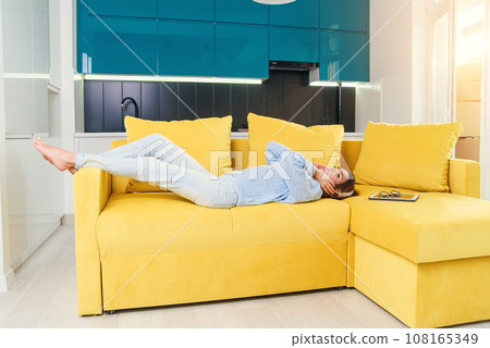 Pretty blonde teenage girl in headphones is listening to music, looking at camera and smiling while lying on her back, legs up, on sofa at home Pretty blonde teenage girl in headphones is listening to music, looking at camera and smiling while lying on her back, legs up, on sofa at home 108165349