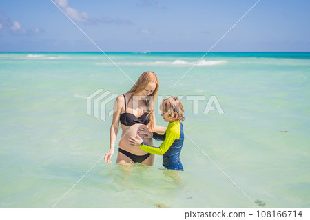 A radiant pregnant mother and her excited son share a tender moment on a serene, snow-white beach, celebrating family love amidst nature's beauty 108166714