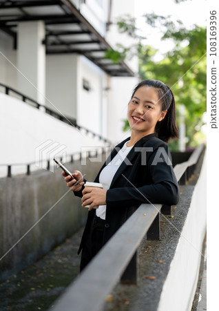 A beautiful Asian businesswoman stands outdoors with her phone and a coffee cup in her hands. A beautiful Asian businesswoman stands outdoors with her phone and a coffee cup in her hands. 108169396
