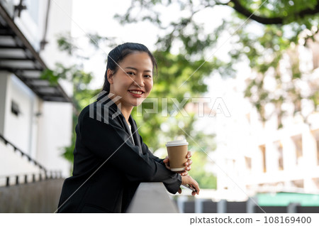 A cheerful and beautiful Asian businesswoman is relaxing outdoors during her coffee break. 108169400