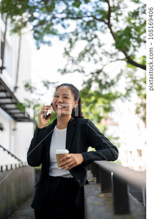 A cheerful Asian businesswoman is enjoying talking on the phone while taking a coffee break outdoors A cheerful Asian businesswoman is enjoying talking on the phone while taking a coffee break outdoors 108169406