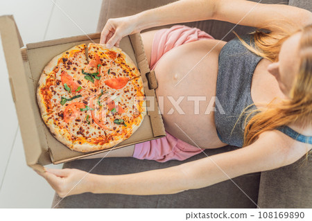 A pregnant woman enjoys a slice of pizza, savoring a moment of indulgence while satisfying her craving for a delightful, comforting treat. Excited Pregnant Young Lady Enjoying Pizza Holding Biting 108169890