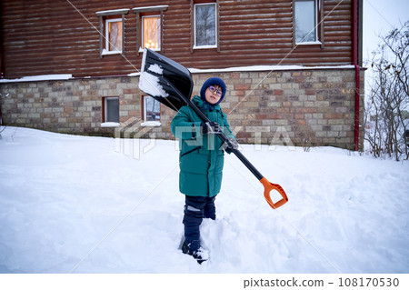 A boy in a green down jacket with a shovel stands in the yard in winter. A boy in a green down jacket with a shovel stands in the yard in winter. 108170530