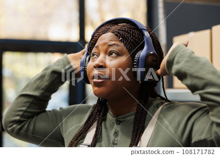 Happy manager with headphones dancing and having fun, working at customers orders preparing packages for shipping in storehouse. Stockroom employee in industrial overall listening music Happy manager with headphones dancing and having fun, working at customers orders preparing packages for shipping in storehouse. Stockroom employee in industrial overall listening music 108171782