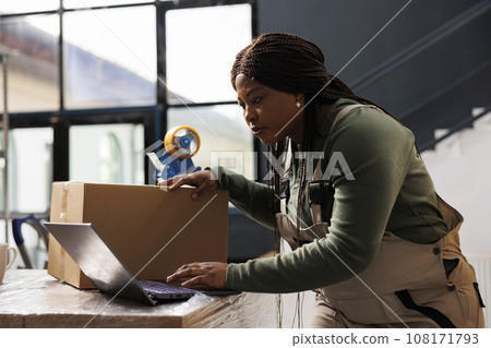 Warehouse manager checking online orders on laptop computer, analyzing shipping details while preparing packages. African american worker analyzing merchandise logistics in storage room 108171793