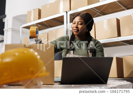 Warehouse supervisor working at clients orders preparing packages for delivery, checking shipping details on laptop computer. African american employee analyzing goods quality control report 108171795