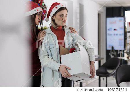Asian employee upset by company manager leaving Christmas decorated office after deciding to quit. Team leader holding desk belongings box, bidding farewell to assistant before starting another job 108171814