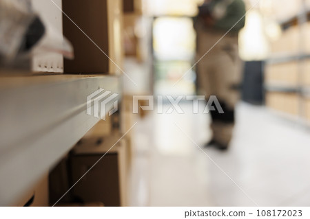 Selective focus of metallic shelves full with carton boxes, in background employee working at customers order preparing packages for delivery in storehouse. Stockroom employee doing products inventory Selective focus of metallic shelves full with carton boxes, in background employee working at customers order preparing packages for delivery in storehouse. Stockroom employee doing products inventory 108172023