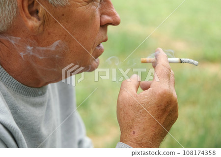 Senior man lighting up a cigarette outdoors against green nature background. Old thoughtful man smoking, looking aside. Close Up Face. Relaxed pensioner enjoy weekend rest outside. Unhealthy habits 108174358