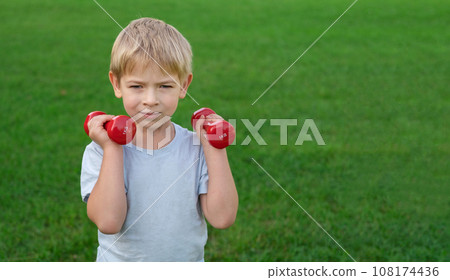 Sporty little boy lifting dumbbells on a green lawn. Joyful toddler doing exercise against nature background outdoors. Happy Child holding red dumbbells, looking at camera. Sport for kids concept. Sporty little boy lifting dumbbells on a green lawn. Joyful toddler doing exercise against nature background outdoors. Happy Child holding red dumbbells, looking at camera. Sport for kids concept. 108174436