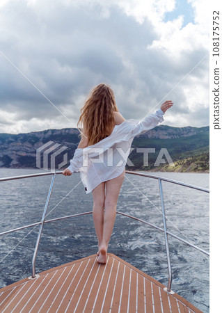 Woman on a yacht. Happy model in a swimsuit posing on a yacht against a blue sky with clouds and mountains Woman on a yacht. Happy model in a swimsuit posing on a yacht against a blue sky with clouds and mountains 108175752