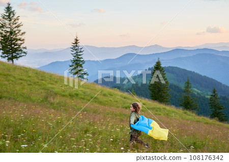 Happy Ukrainian running in Carpathian mountains. Little kid smiling and flattering Ukrainian flag on background of hills at sunset. Happy Ukrainian running in Carpathian mountains. Little kid smiling and flattering Ukrainian flag on background of hills at sunset. 108176342