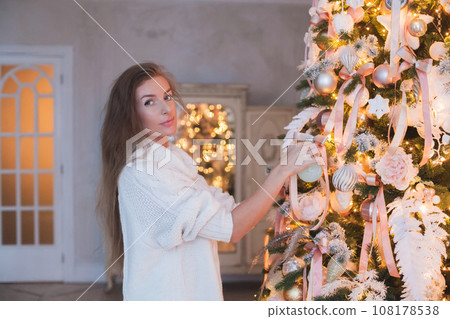 Beautiful woman in white sweater standing with Xmas tree and Christmas decoration at home 108178538