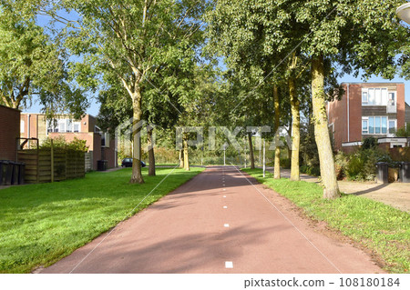 a tree lined street with houses in the background and green grass on both sides, along a path that leads to an apartment complex 108180184
