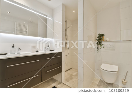 a modern bathroom with white and brown cabinets, toiletries, sink, mirror and shower stall in the corner 108180229