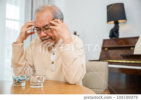 Asian senior old man grey haired sad sitting in chair hand hold head with many medicines and pills for taking, elderly man sick have headache looking pill glass on at home, Elderly healthcare Asian senior old man grey haired sad sitting in chair hand hold head with many medicines and pills for taking, elderly man sick have headache looking pill glass on at home, Elderly healthcare 108180979
