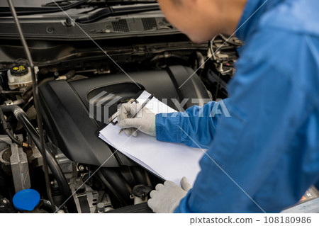 Close-up shot of an Asian mechanic's hand, in blue workwear, writing notes on a paper while working on a car in an auto repair shop. 108180986