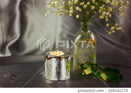 A burning candle in metal holder on silver silk tablecloth. Luxurious stylish decor for modern interior. A little vase with rural small wild white flowers, green heart-shaped glasses on cosmetic table A burning candle in metal holder on silver silk tablecloth. Luxurious stylish decor for modern interior. A little vase with rural small wild white flowers, green heart-shaped glasses on cosmetic table 108182270