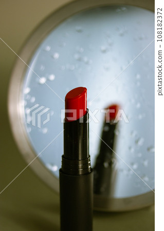 Women's makeup. Red lipstick in tube next to a round mirror with reflection on water drops on a makeup table. Cosmetic still life in dark key. Decorative cosmetics sample on vertical grey background. Women's makeup. Red lipstick in tube next to a round mirror with reflection on water drops on a makeup table. Cosmetic still life in dark key. Decorative cosmetics sample on vertical grey background. 108182372