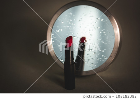 Women's makeup. Red lipstick in tube next to a round mirror with reflection on water drops on a makeup table. Cosmetic still life in dark key. Decorative cosmetics sample on vertical grey background. Women's makeup. Red lipstick in tube next to a round mirror with reflection on water drops on a makeup table. Cosmetic still life in dark key. Decorative cosmetics sample on vertical grey background. 108182382